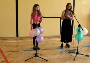 Uczennice na sali gimnastycznej stoją przy mikrofonach i śpiewają przygotowane przez siebie utwory w ramach pokazu szkolnych talentów.